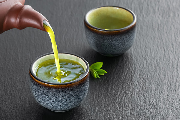 Hot green tea is poured from the teapot into the blue bowl, darck stjne table. Tea leaves next to the cup. Close-up, tea ceremony, minimalism, copy space for text.